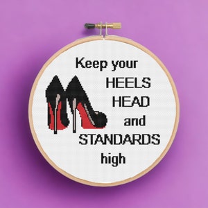 May include: A cross-stitch embroidery hoop featuring black high heels with red soles. The text reads "Keep your HEELS HEAD and STANDARDS high" in black block letters. The hoop is set against a purple background.