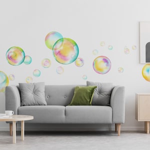 Puede incluir: Un sofá gris con cojines verdes se encuentra frente a una pared blanca con calcomanías de burbujas de acuarela. Una mesa de café de madera con libros se encuentra frente al sofá. Un gabinete de madera con estantes se encuentra en el lado derecho de la imagen.
