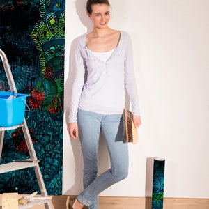 May include: A woman stands near a wall with a colourful patterned wallpaper. She is holding a paint roller and a bucket of paint. The wallpaper is a dark blue and green colour with a floral pattern.
