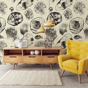 Op de afbeelding: Een gele fauteuil met houten poten staat voor een houten kast met drie lades. De kast staat voor een muur met een zwart-wit behangpatroon van boomringen.