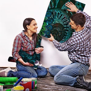 May include: A couple working on a home improvement project. The man is holding up a large, colourful, abstract print while the woman sits on the floor with paint rollers and paint cans.
