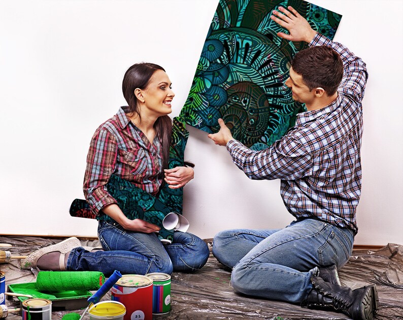 May include: A couple working on a home improvement project. The man is holding up a large, colourful, abstract print while the woman sits on the floor with paint rollers and paint cans.