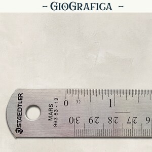 May include: A stainless steel ruler with black markings and the text "STAEDTLER MARS 963 53-12". The ruler measures up to 2 inches and 30/32 of an inch. The word "GIOGRAFICA" is at the top.