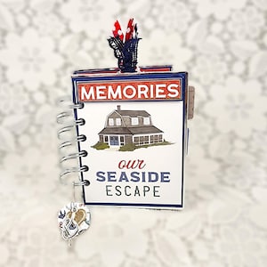 May include: A small spiral-bound scrapbook with a white cover featuring a brown house illustration and the text MEMORIES our SEASIDE ESCAPE. The book has a silver metal coil binding and a decorative charm with a heart and anchor hanging from it.