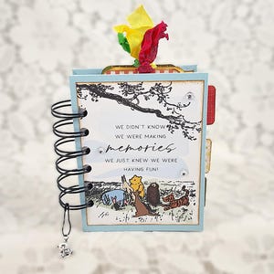 May include: A small, spiral-bound scrapbook with a light blue cover. The cover features a quote about memories and illustrations of Winnie the Pooh characters. A charm hangs from the spiral binding, and colorful ribbons adorn the top.