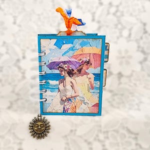 May include: A small, blue-framed journal with a spiral binding and a beach scene on the cover. The cover art features two women with umbrellas. A sun charm hangs from the journal. A colorful ribbon is attached to the top.