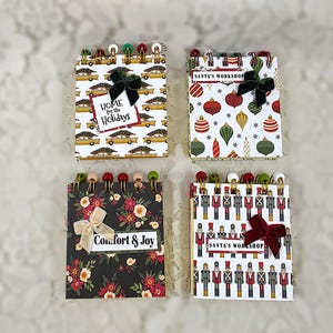 May include: Four square spiral-bound notebooks featuring holiday themes. One shows cars with trees and says Home for the Holidays, one has ornaments and says Santa's Workshop, one has floral patterns and says Comfort & Joy, and one has nutcrackers.