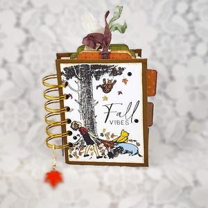 May include: A small spiral-bound notebook with a brown cover featuring a printed illustration of characters in a forest with falling leaves and the text Fall VIBES. The notebook includes gold wire binding, a hanging red leaf charm, and fabric ribbon accents.