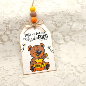 May include: A wooden tag with a honey bear illustration and the text "Taste and See that the Lord is GOOD, Psalm 34:8". The tag is decorated with yellow and brown beads.