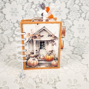 May include: A small, square, spiral-bound scrapbook with an orange frame. The cover features an illustration of a house porch decorated with orange pumpkins. A silver maple leaf charm hangs from the white plastic coil, and a fabric ribbon is tied at the top.