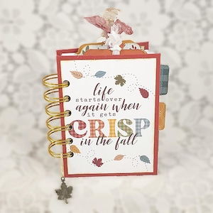 May include: A small spiral-bound notebook with a white cover featuring the text "life starts over again when it gets CRISP in the fall" in brown and plaid lettering. The cover is decorated with small colorful autumn leaves and a metal leaf charm.