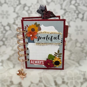 May include: A small spiral-bound notebook with a dark red cover and copper-colored wire binding. The front features a grid-patterned panel with the text "grateful" and "ALWAYS", decorated with paper sunflowers, leaves, and a hanging metal charm.
