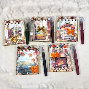 May include: Five small spiral-bound notebooks featuring autumn-themed cover art, including scenes of a window, a basket of apples, a landscape, and a fireplace. Each notebook is adorned with a small velvet bow and includes a matching ballpoint pen.