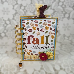 May include: A spiral-bound notebook with a white cover featuring a colorful pattern of autumn leaves and the text "fall-lelujah!". The notebook is decorated with fabric flowers in red and yellow, and a small acorn charm hangs from the gold wire coil.