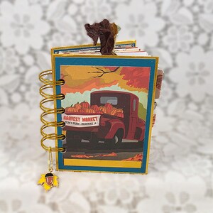 May include: A small spiral-bound notebook featuring a cover illustration of a red pickup truck loaded with pumpkins and the text Harvest Market Tom's Farm Highway 14. The notebook has gold wire binding and a small dangling acorn charm at the bottom.