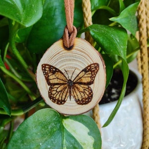 May include: A round wooden pendant with a brown leather cord. The pendant features a detailed butterfly design burned into the wood. The butterfly has brown and tan wings. The pendant hangs in front of green leaves.