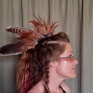 May include: A woman with long, brown hair with pink highlights wears a brown and black feather headdress. She has a braided hairstyle and is wearing makeup with a brown and black design around her eye.