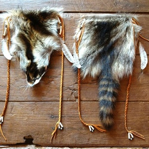 May include: Two brown leather pouches with raccoon fur and tails. The pouches have white feathers and brown leather straps with white beads.