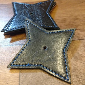 May include: Two black leather ninja stars with blue stitching. The stars are stacked on top of each other. The top star has the word "Ninja" written on it.