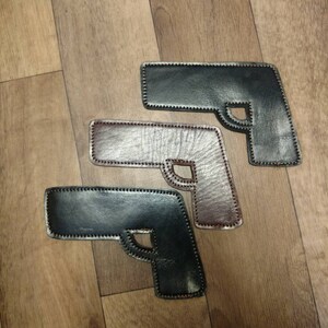 May include: Three leather gun holsters in different colors: black, brown, and dark brown. The holsters are shaped like a gun and have a cut-out for the trigger.