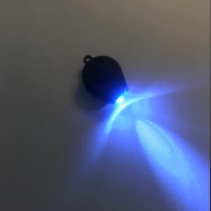 Puede incluir: Una linterna negra con forma de lágrima con una luz azul brillante. La luz emana de la parte inferior de la linterna, creando un efecto de explosión estelar. El llavero tiene un pequeño lazo en la parte superior para sujetarlo.