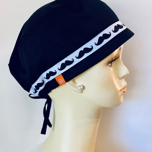 May include: Black scrub hat with a white ribbon trim featuring a repeating pattern of black mustaches.