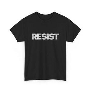 Puede incluir: Camiseta negra con la palabra "RESIST" impresa en blanco. La palabra está compuesta por texto más pequeño, creando un efecto texturizado. La camiseta tiene mangas cortas y cuello redondo.