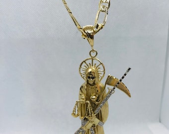 May include: A gold chain necklace with a pendant featuring a detailed depiction of Santa Muerte, a skeletal figure holding a rifle, scales, and a scythe. The pendant is adorned with small, clear stones.