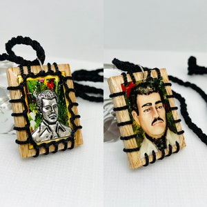 May include: Two wooden framed pendants with black cord necklaces. Each pendant features a portrait of a man with a black and white image inside. The pendants are decorated with black string and gold trim.