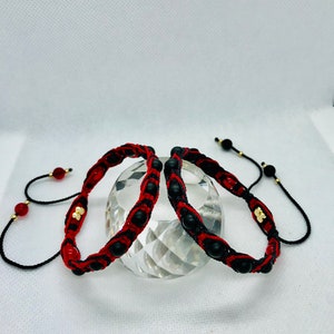 May include: Three black and red braided bracelets with black beads and gold accents. The bracelets are tied together with black cords.