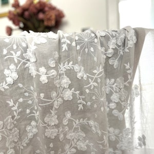 以下が含まれることがあります： 繊細な花の刺繍模様が施された白い綿生地。刺繍は、透き通るような白い背景に白い花と葉が描かれています。