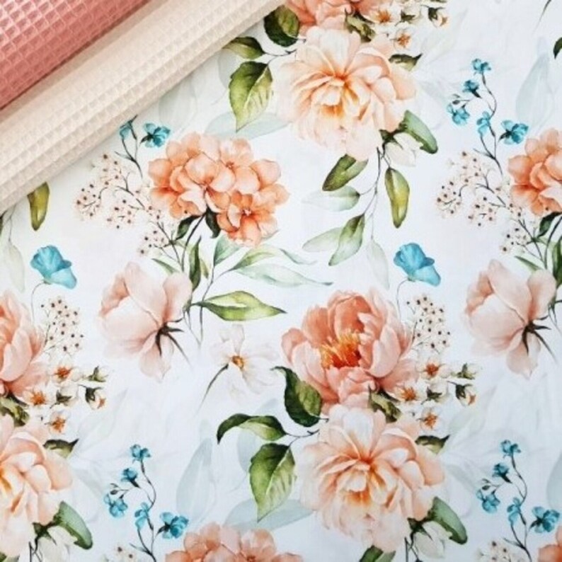 May include: A white fabric with a repeating pattern of peach and white flowers, green leaves, and small blue flowers.