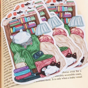 May include: Three illustrated stickers featuring a cat sleeping in a green armchair with a book shelf behind it. The cat is wearing a red scarf and there is a lamp on the table next to the chair. The stickers are on a book with text about the Atlantic colonies.