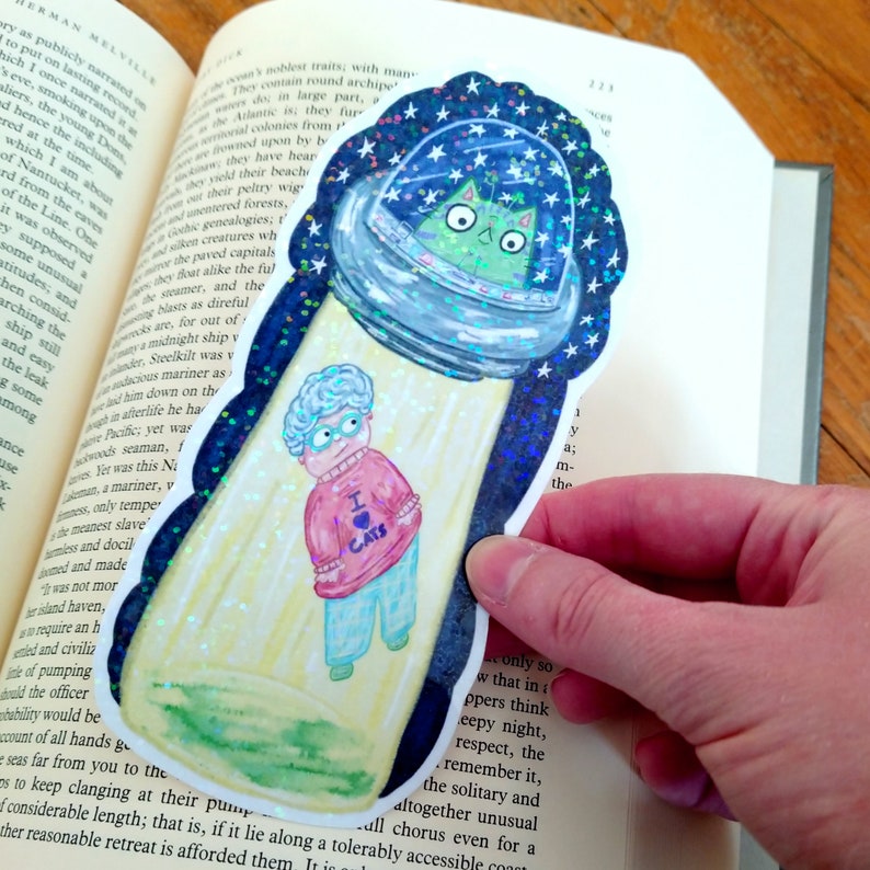 May include: A colorful sticker featuring a cartoon grandma wearing a pink sweater with the words "I love cats" on it. She is standing in front of a flying saucer with a green cat inside. The sticker has a glittery background.