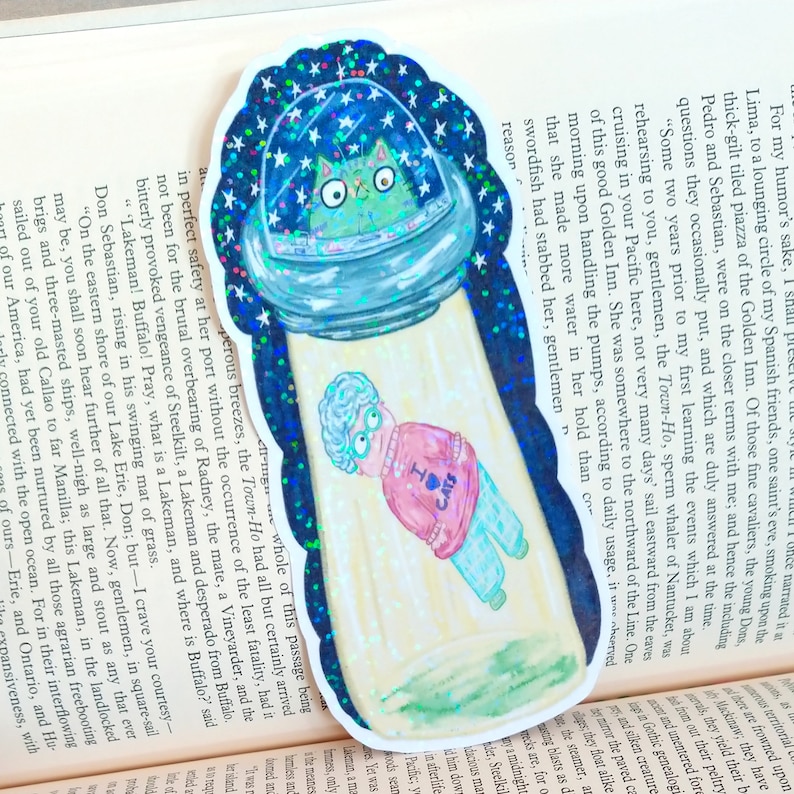 May include: A glittery sticker featuring a green cat in a space helmet floating above a woman wearing a pink sweater with the text "I &hearts; Cats" on it. The sticker is set against a background of a starry night sky and green grass.