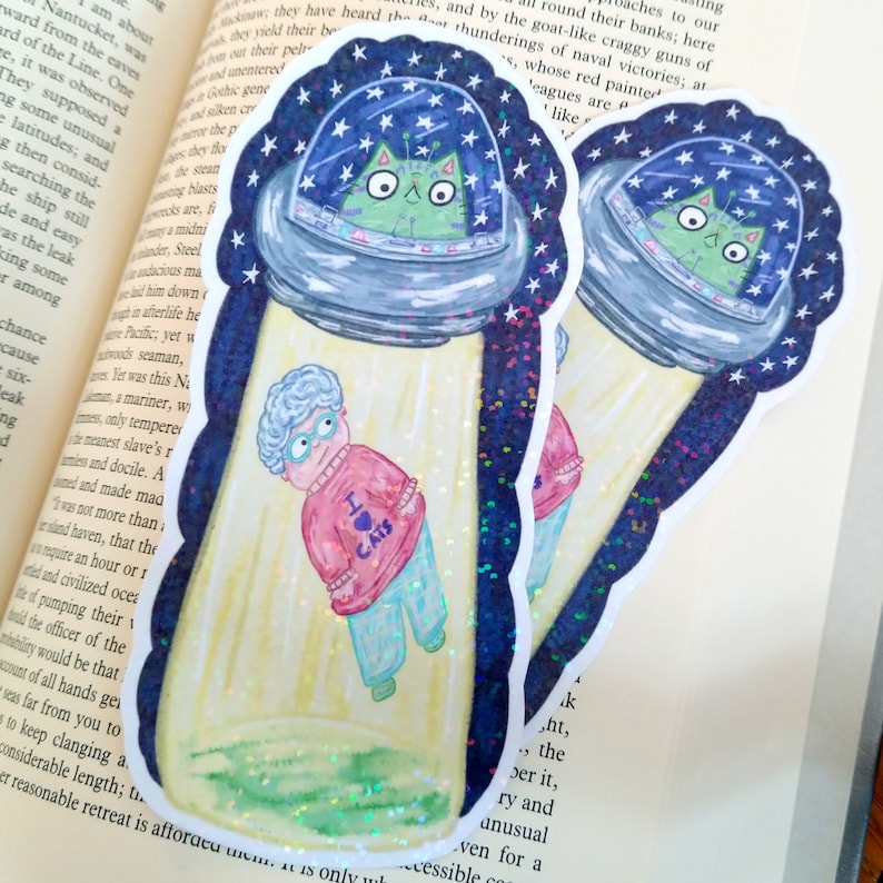May include: Two holographic stickers featuring a cartoon cat in a flying saucer, with a beam of light shining down on a woman wearing a pink sweater with the words "I love cats" on it. The stickers are on a book.