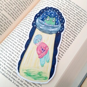 May include: A watercolor illustration of a green cat in a flying saucer with a starry night sky background. A woman wearing a pink sweater with the words "I love cats" is standing in front of the saucer. The illustration is printed on a bookmark.