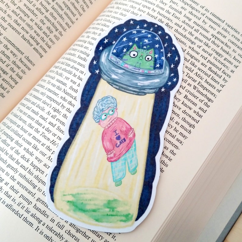 May include: A watercolor illustration of a green cat in a flying saucer with a starry night sky background. A woman wearing a pink sweater with the words "I love cats" is standing in front of the saucer. The illustration is printed on a bookmark.