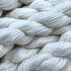 以下が含まれることがあります： 編み込まれた質感の、太いオフホワイトの糸のクローズアップ。糸には、きらめきを加えるために、微妙な銀色のメタリック糸が織り込まれています。この画像は、繊維の質感と詳細を強調しており、クラフトプロジェクトに適しています。