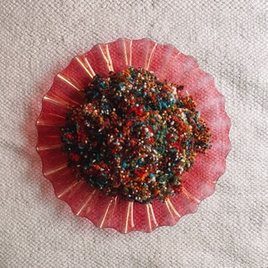 Può includere: Una vista dall'alto di un piatto di vetro rosso con bordo a balze, pieno di filato colorato. Il filato è un mix di rosso, arancione, blu, verde e bianco, creando un aspetto strutturato e maculato. Il piatto è su una superficie neutra e strutturata.