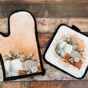 May include: A set of two oven mitts with a fall theme. The mitts feature a white pumpkin with a brown stem and a brown and white knitted pumpkin with a brown bow. The mitts have a white background with brown and orange accents. The text "Silver Harvest" is printed on the mitts.