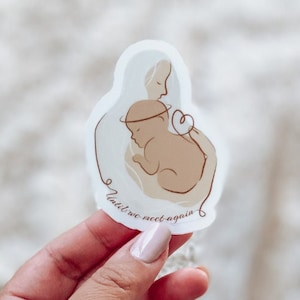 May include: A white sticker with a drawing of a mother holding a baby. The baby has a halo above its head and the mother has a heart above her head. The text "Until we meet again" is written in a cursive font around the drawing.