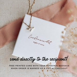May include: A gold necklace with a heart-shaped pendant with wings and a clear stone, displayed on a white box. The box has the text "Eve Amara light & love" and the words "send directly to the recipient!" are also visible.