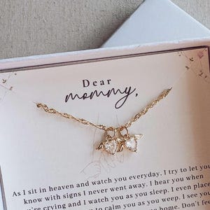 May include: A gold necklace with heart-shaped pendants, displayed in a white gift box with the words "Dear mommy," printed on a card. The box is on a light-coloured surface.