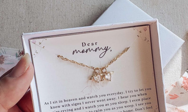 May include: A gold necklace with heart-shaped pendants, displayed in a white gift box with the words "Dear mommy," printed on a card. The box is on a light-coloured surface.