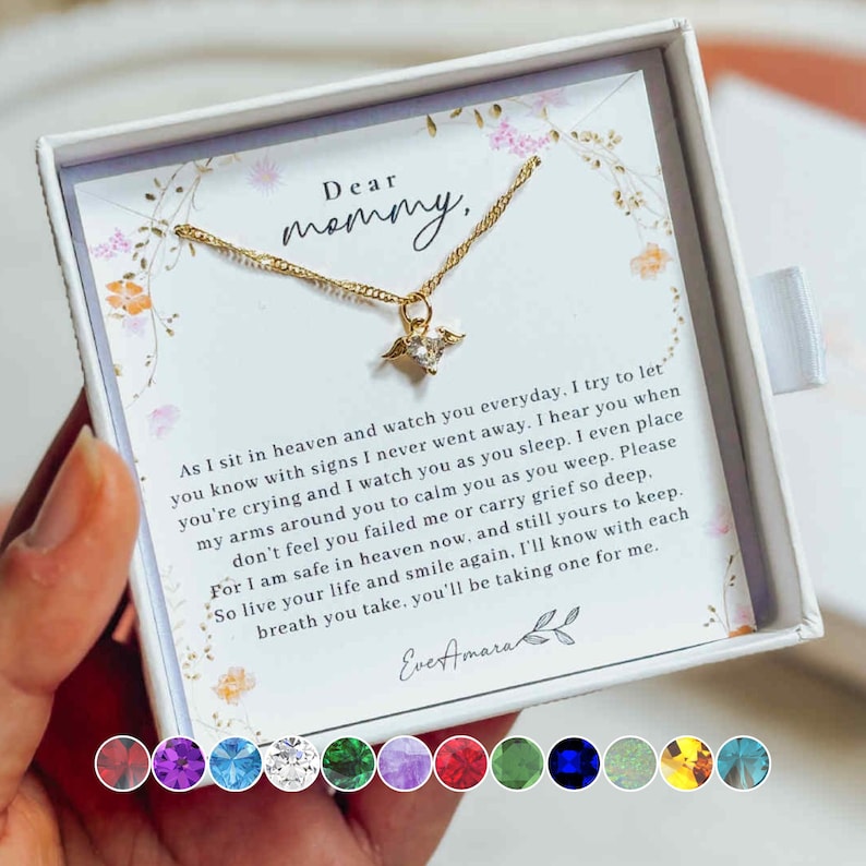 May include: A gold necklace with a winged pendant, displayed in a white box with floral accents. The box contains a card with the words "Dear mommy" and a touching message. The necklace is a thoughtful gift.