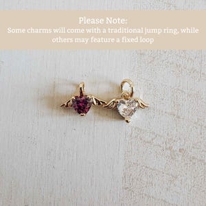 May include: Two gold heart-shaped charms with wings. One has a deep red gemstone, the other a clear gemstone. The charms are on a light surface. Text at the top says "Please Note: Some charms will come with a traditional jump ring, while others may feature a fixed loop."
