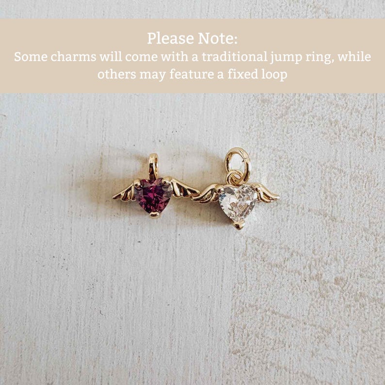 May include: Two gold heart-shaped charms with wings. One has a deep red gemstone, the other a clear gemstone. The charms are on a light surface. Text at the top says "Please Note: Some charms will come with a traditional jump ring, while others may feature a fixed loop."