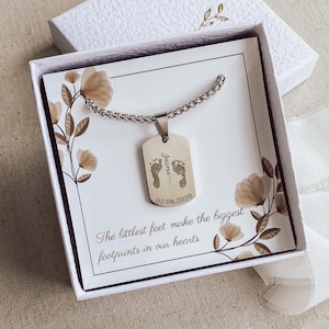 May include: A silver necklace with a dog tag pendant engraved with the name "Jaime" and the date "02.08.2020". The pendant also features a pair of tiny footprints. The necklace is displayed in a white box with a floral design and the text "The littlest feet make the biggest footprints in our hearts".