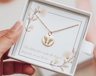 May include: Gold necklace with a round pendant featuring a baby's footprint. The necklace comes in a white box with a floral design and the text "The littlest feet make the biggest footprints in our hearts."